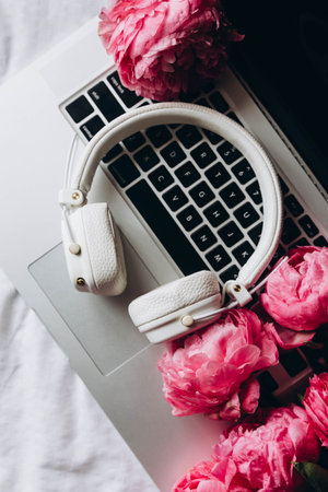 White headphones, laptop and pink peony flowers on white sheets in bed, concept of remote work, music creative. Flat lay, top view spring minimalist home workspace.の写真素材