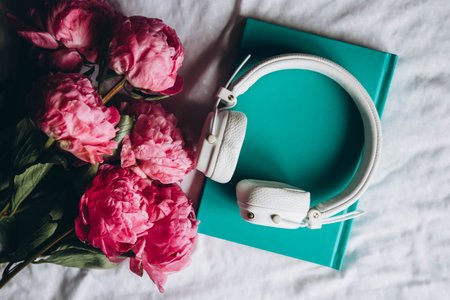 Book, white headphones and a bouquet of spring pink peony flowers on the bed, top view. Audiobook, reading and relaxation concept.の写真素材