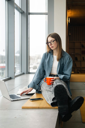 Stylish young woman with glasses uses a laptop, sits in a university library.の写真素材