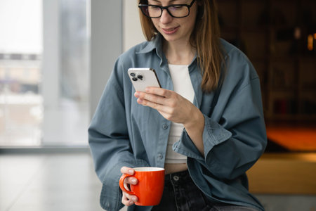 A woman is using a smartphone and holding a cup while sitting by the window.