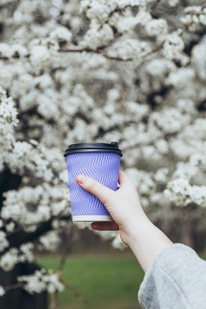 Paper cup in the hand of a girl on a background of white blooming spring tree.