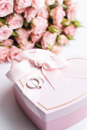 A heart-shaped gift box and a pair of wedding rings on a background of flowers.の写真素材