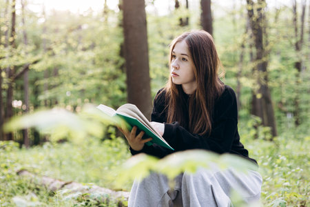Teenage girl reading a book while sitting on a log in the forest, copy space.の写真素材