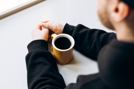 Man with hands clasped near yellow mug of filter coffee on bright tabletop.の写真素材