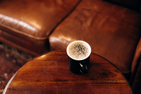 Black coffee mug on wooden table near leather sofa with copy space and daylight.