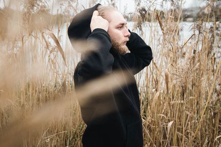 A young bearded man in a black hoodie in the reeds near the river.の写真素材