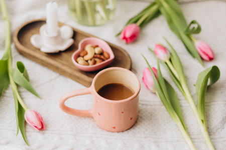 Close-up of a coffee cup with pink tulips, candle, and plate of nuts at home.