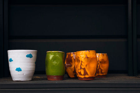 Colorful handmade ceramic cups displayed on a dark shelf with ample copy space.