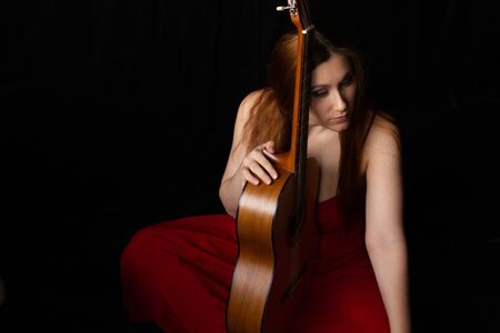 girl in a red dress sits on the floor near the guitar on a black backgroundの写真素材