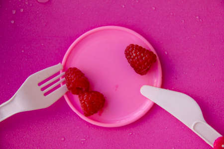 three raspberry berries lie on a small pink plate, next to a plastic fork and and a knife