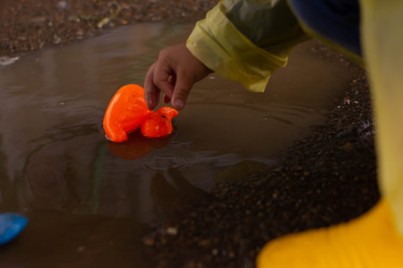 Beautiful girl in denim suit and yellow rubber boots plays with plastic duck and whale in a puddleの写真素材