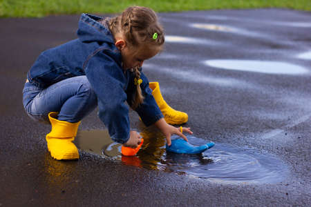 Beautiful girl in denim suit and yellow rubber boots plays with plastic duck and whale in a puddleの写真素材