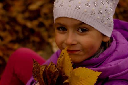 portrait of a pretty girl in a purple jacket and white hat in the fall. She holds yellow leaves and...