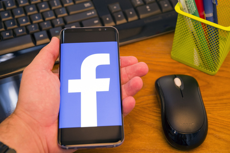 PIATRA NEAMT, ROMANIA - JULY 30, 2018: Hand holds a Samsung S8+ with Facebook logo on the screen, office background.のeditorial素材