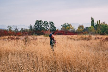 Beautiful woman is walking in the park in cloudy weather at autumn. Red-haired female in a blue hat and coat walks the field at fall looking to the skyの写真素材