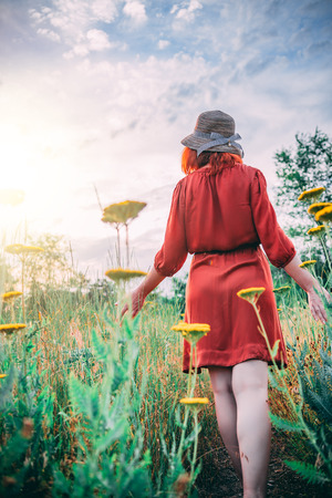 Girl on dandelion on green field. A lonely beautiful  woman  in a red dress is walking and enjoying the greenery and dandelion.の写真素材