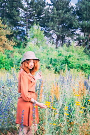 Girl on dandelion on green field. A lonely beautiful girl in a red dress with hat  is walking and enjoying the greenery and dandelion.の写真素材