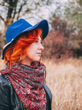 Charming woman is walking in the park in cloudy weather at autumn. A red-haired female in a blue hat walks the field in fall rainy weather.の写真素材