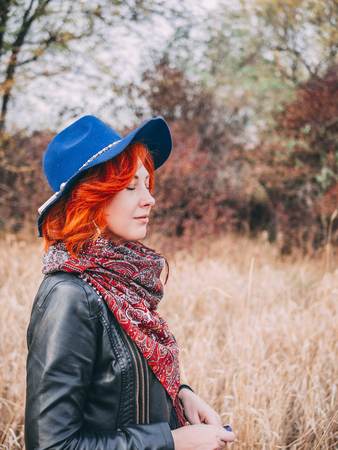 Awesome woman is walking in the park in cloudy weather fall. A red-haired female in a blue hat walks the field in autumn rainy weather.の写真素材