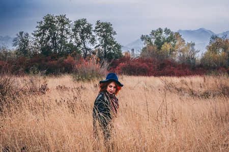 Pretty woman is walking in the park in cloudy weather at autumn. A red-haired female in a blue hat and coat walks the field at fallの写真素材