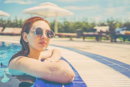 Woman looks out of the pool, hanging on the railing on the background of sun beds with umbrellas. Female swimming in the pool. Young woman relaxing in the water . Tonedの写真素材