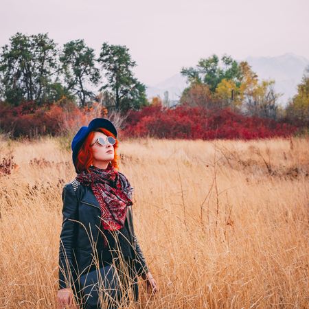 Awesome woman is walking in the park in cloudy weather at autumn. Red-haired female in a blue hat, coat and sunglasses walks the field at fall looking to the sky. Copy space for text. Crop 1x1の写真素材