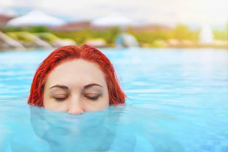 Beautiful sexy female relaxing In swimming pool water. Girl with healthy tanned skin, gorgeous face and enjoying sun on hot summer day. Half head above water stalking. Relax concept with copy spaceの写真素材