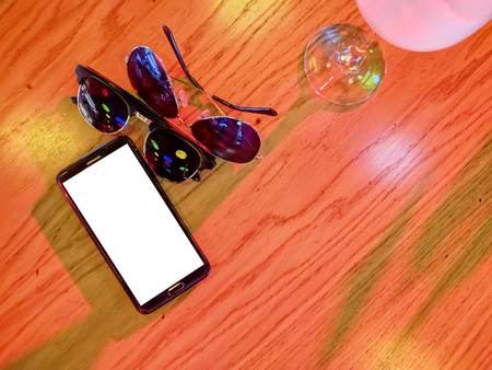 Phone and a couple glasses. Modern black smart phone with white isolated display for mockup in wooden table. Cocktail, glasses.の写真素材