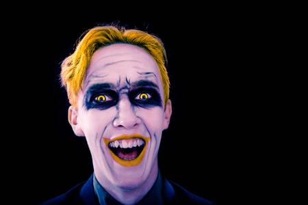 Young man with white face, yellow eyes hair and lips celebrates halloween. Smiling clown on the black background. Halloween party. Copy space for text.の写真素材