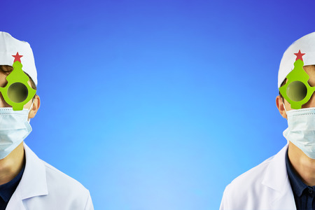 Two cool young doctors in a white hat and a medical mask in a Bathrobe and black glasses with a festive Christmas tree on them on satureated blue background with copy space for textの写真素材