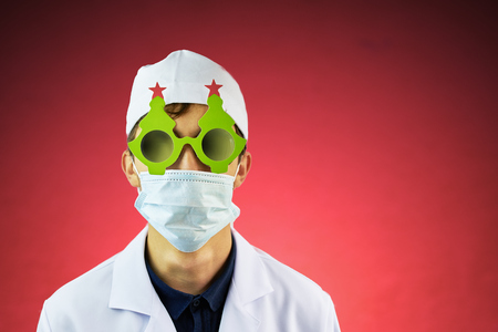 Young doctor in a white hat and a medical mask in a Bathrobe and black glasses with a festive Christmas tree on them. Holiday costume with copyspace for textの写真素材