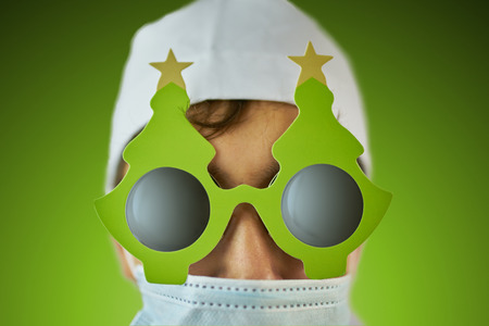 Young doctor in a white hat and a medical mask in a coat and black glasses with a holiday Christmas tree on them on saturated green background in the centerの写真素材