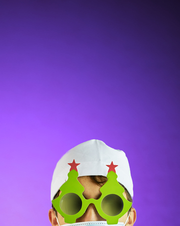 Doctor in a white hat and a medical mask in a Bathrobe and black glasses with a festive Christmas tree on them on purple background with vertical copyspaceの写真素材