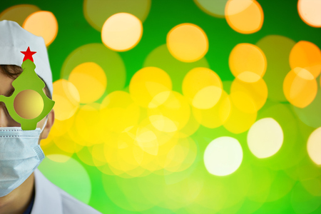 Crazy medic in Christmas tree glasses and white coat and hospital mask on green background with bright yellow lights. Copy space for text. Merry Christmas Doctorの写真素材