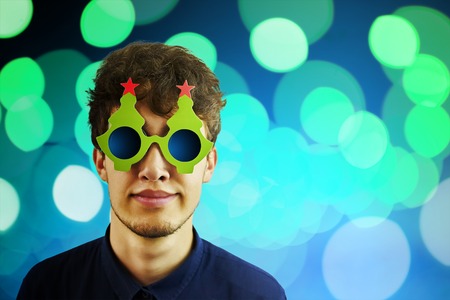 Portrait of a man in Christmas glasses. Portrait of cheerful, attractive man on blue sparkles background with copy space for text, look over glasses make wide smile. Copyspace for textの写真素材
