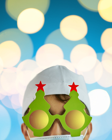 Crazy physician in Christmas tree glasses with red stars and white coat on blue background with bright lights with copy space for text. Vertical crop 4x5. Merry Christmas Doctorの写真素材