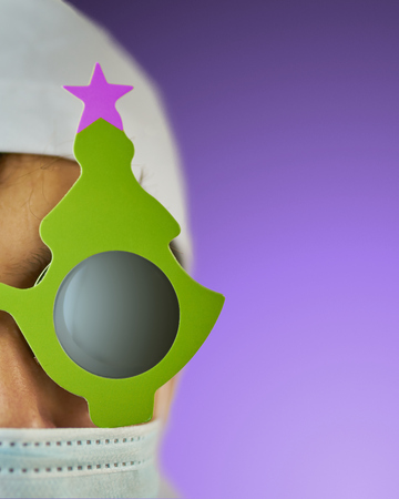 Young doctor in a white hat and a medical mask in a Bathrobe and black glasses with a festive Christmas tree on them. Holiday costume. Verticalの写真素材