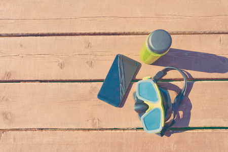 Swimming mask, bottle of smoothie and  smartphone on wooden table on sunny day on vacationの写真素材