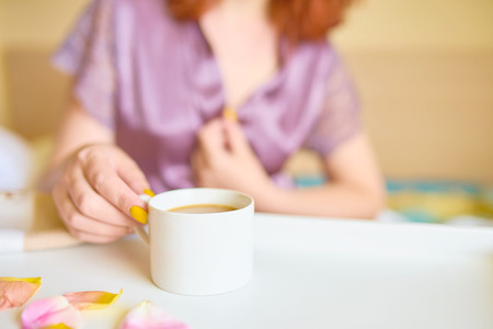 Pretty cute woman in a purple silk robe drinking coffee early in the morning in bed. White cup of latte on the tray. Breakfast concept with copy spaceの写真素材