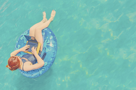Young pretty woman freelancer is floating on the sea or in the pool in a swimming circle. A girl is relaxing on the ocean or in the dug-out pool on the inflatable ring with laptop and copyspaceの写真素材