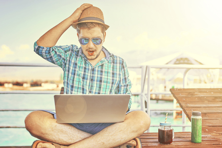 A man on vacation, sits on the pier and works. Concept of workaholic. Freelancer is working on time. The shocked face of the worker, in the computer.の写真素材