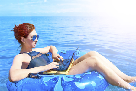 Beautiful young woman with an inflatable ring and a laptop relaxing in the sea. Workaholic conceptの写真素材