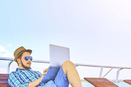 Freelancer guy lies on a sunbed and works on a laptop with sunglasses and hat. Hipster workaholic concept. Businessman Working Summer Beach Relaxation Concept.の写真素材