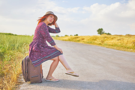 Very pretty woman in summer dress and hat hitchhiking with suitcase on the road. Beauty woman sitting on Luggage waiting for bus. Hitchhiker conceptの写真素材
