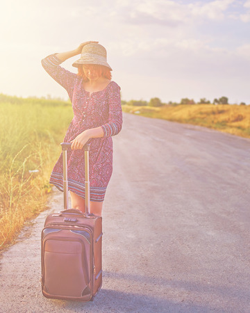Cool female tourist in summer dress and hat hitchhiking with suitcase on the road. Woman stands next to the Luggage waiting for the transport. Travel concept with pink tonedの写真素材