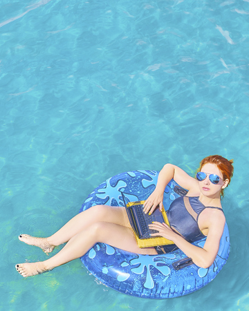 Young pretty woman freelancer is floating on the sea or in the pool in a swimming circle. A girl is relaxing on the ocean or in the dug-out pool on the inflatable ring with laptop. Vertical 4x5 cropの写真素材
