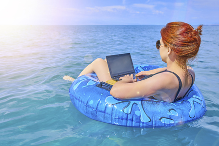 Beautiful young woman with an inflatable ring and a laptop relaxing in the sea and working on the laptop. Freelancer on the vacation. Workaholic conceptの写真素材