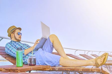 Freelancer guy lies on a sunbed and works on a laptop and drinks smoothie. Hipster workaholic concept. Businessman Working Summer Beach Relaxation Concept.の写真素材