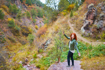 Awesome woman tourist with a backpack admires the mountain rock. Pretty female hiker taking selfie or making live video stream vlog in mountains forest.の写真素材