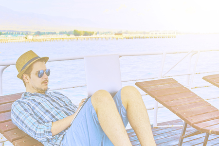 Young businessman resting on piers, enjoying summertime in resort spa near sea while working remotely at laptop computer connected to wireless internet.の写真素材
